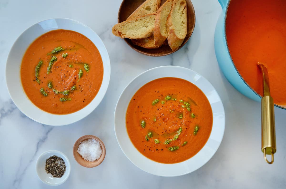 Two white bowls are filled with tomato soup and drizzled with basil pesto. Small bowls of salt, pepper and sliced baguette are nearby.