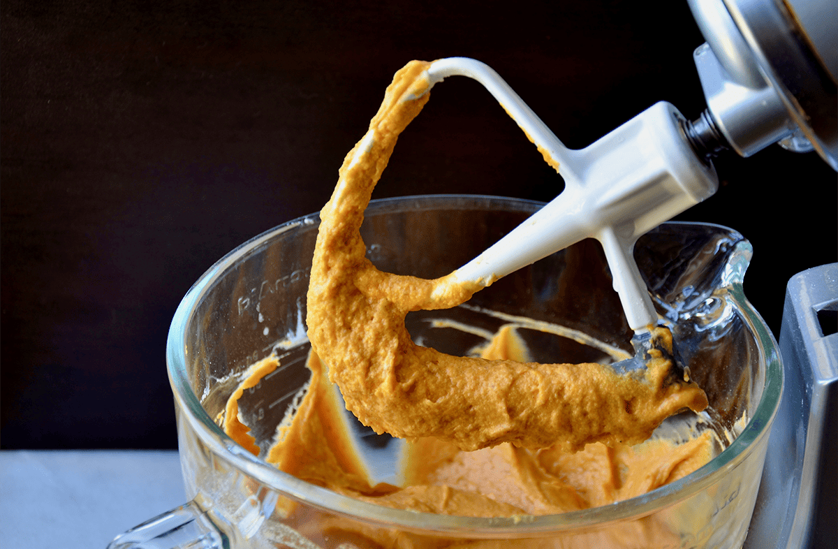 Pumpkin muffin batter in the bowl of a stand mixer with the stand mixer's paddle coated in batter.