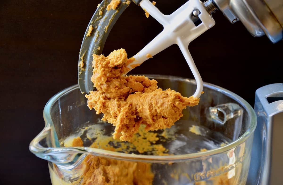 Biscotti dough in the bowl of a stand mixer fitted with a paddle attachment. A gob of dough is stuck to the paddle.