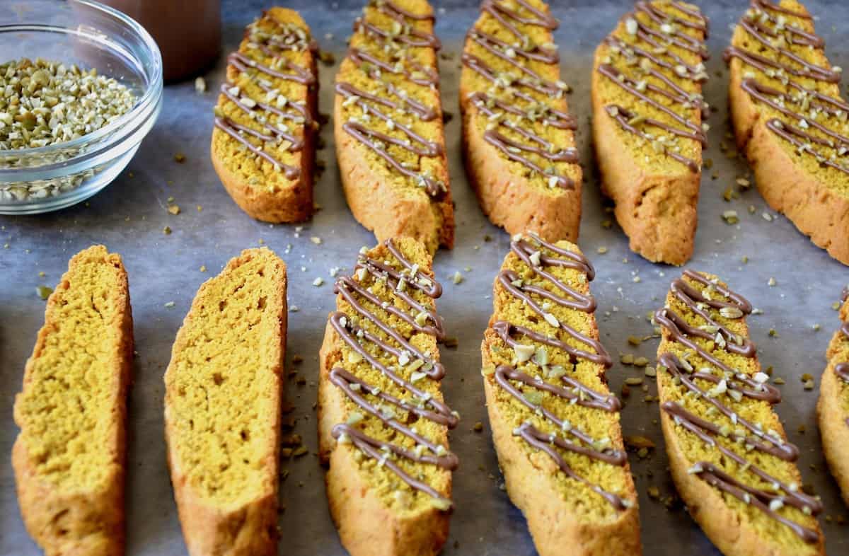 Two rows of pumpkin pie biscotti, with all but two of the biscotti drizzled with melted chocolate and chopped pumpkin seeds. Two small bowls of more chopped pumpkin seeds and melted chocolate sit beside the biscotti.