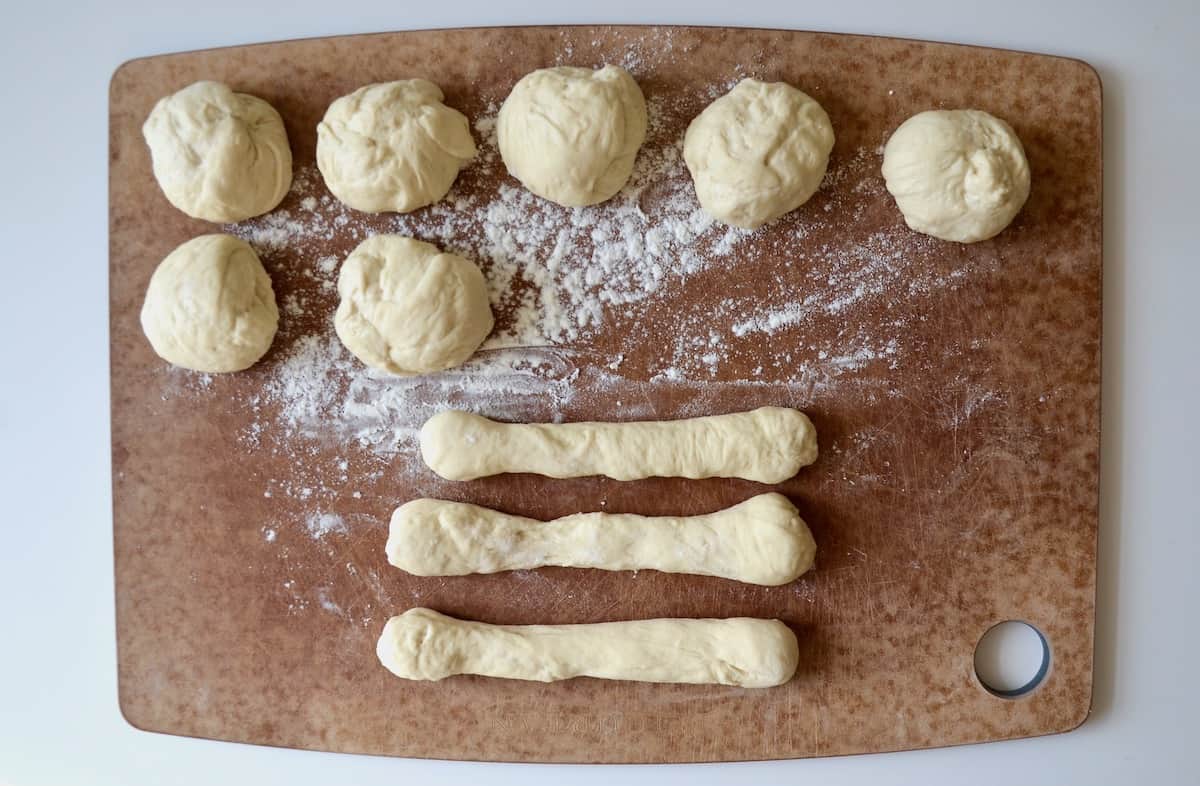 Three pieces of dough rolled out into ropes beside 7 balls of dough on a floured wooden cutting board.