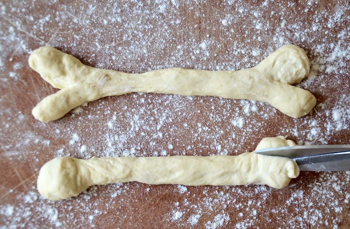 A pair of kitchen shears snipping the end of a rope of dough. A snipped and shaped breadstick bone sits beside it.