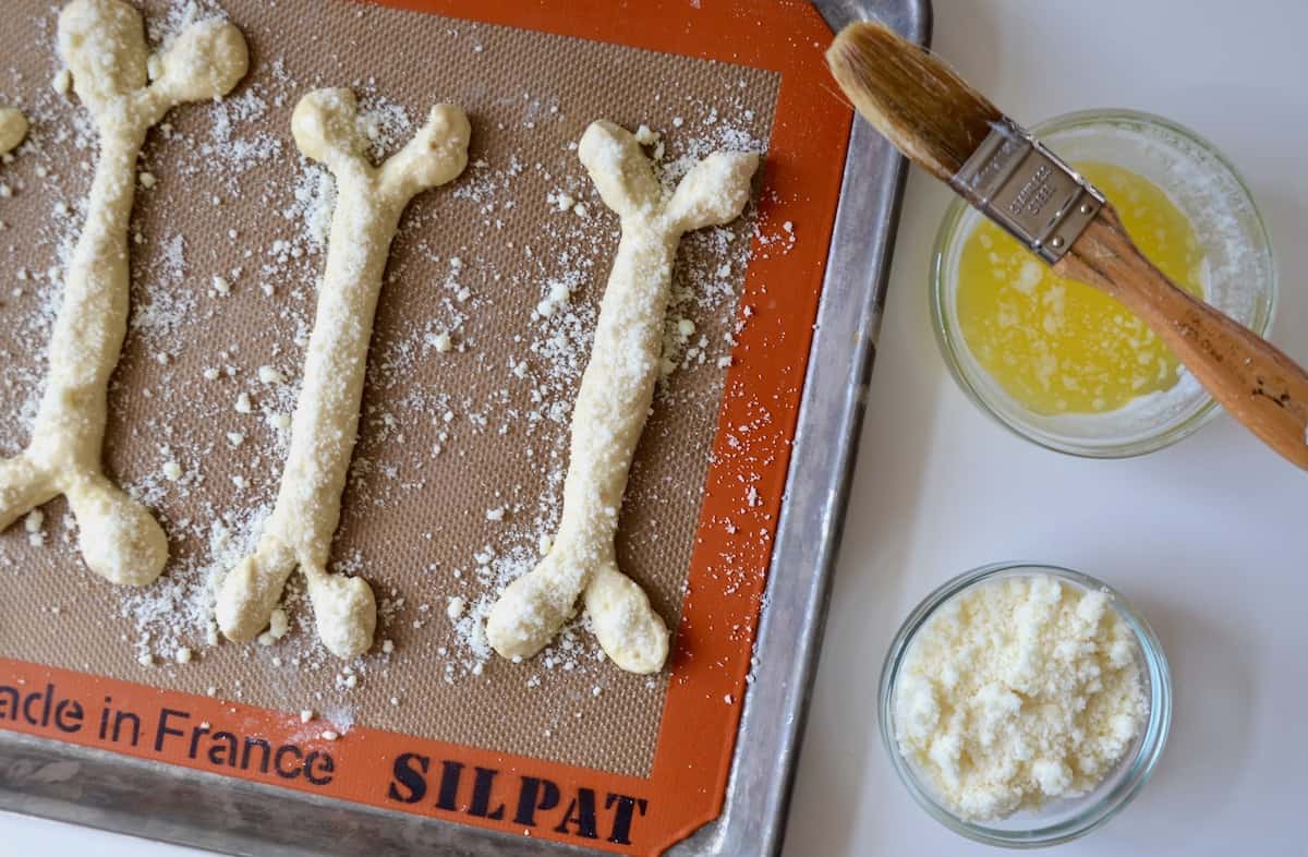 Shaped breadstick bones topped with grated Parmesan cheese on a baking sheet lined with Silpat. Beside the baking sheet sits small bowls of grated cheese and melted butter, with a pastry brush balanced on top of the bowl of butter.