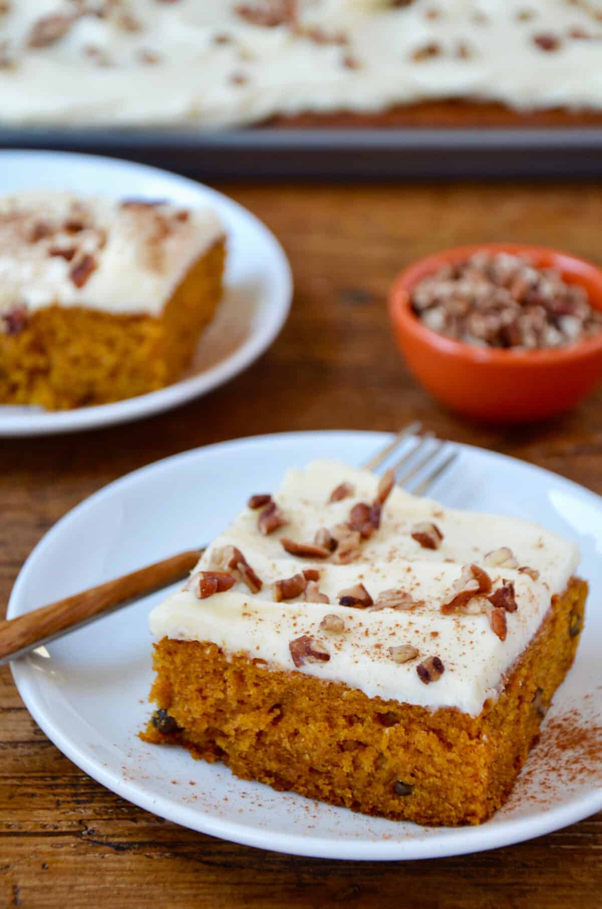 Two white plates hold slices of pumpkin bars with cream cheese frosting, sprinkled with chopped pecans. A red bowl of pecans is in the background.