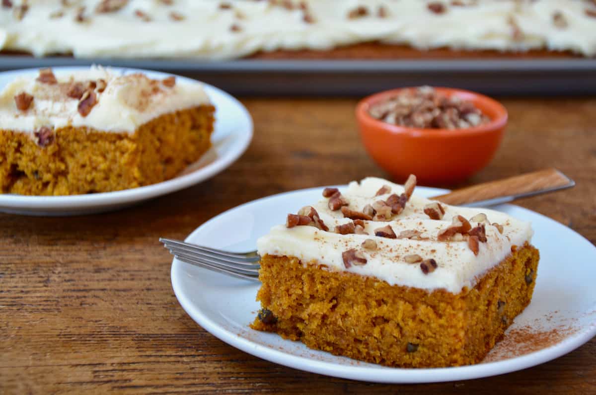 Two white plates hold slices of pumpkin bars with cream cheese frosting, sprinkled with chopped pecans. A red bowl of pecans is in the background.