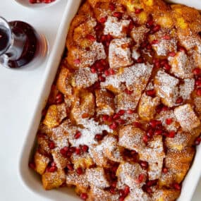 A baking dish of pumpkin French toast casserole with a container of maple syrup and bowl of pomegranate arils beside it.