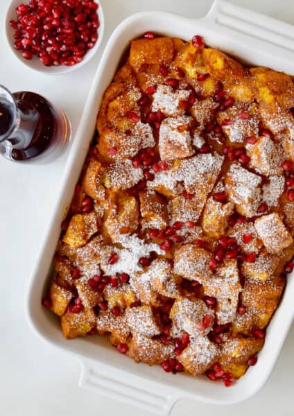 A baking dish of pumpkin French toast casserole with a container of maple syrup and bowl of pomegranate arils beside it.