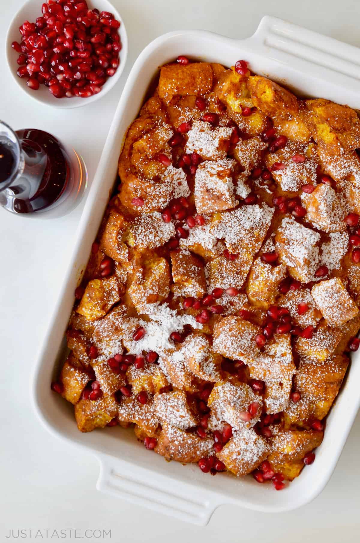 A baking dish of pumpkin French toast casserole with a container of maple syrup and bowl of pomegranate arils beside it.