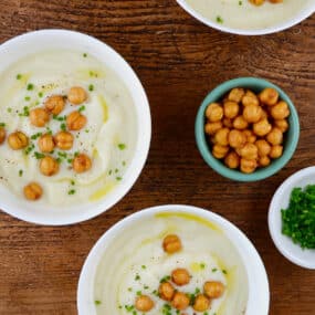 Three bowls of of cauliflower soup topped with crispy chickpeas, minced chives and olive oil. Small bowls of extra crispy chickpeas and minced chives are beside the soup bowls.