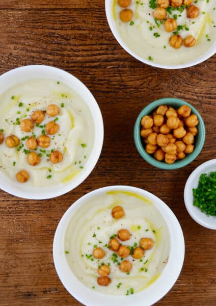 Three bowls of of cauliflower soup topped with crispy chickpeas, minced chives and olive oil. Small bowls of extra crispy chickpeas and minced chives are beside the soup bowls.