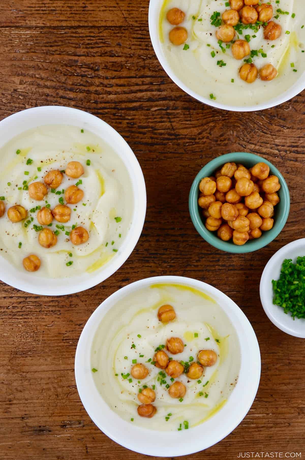 Three bowls of of cauliflower soup topped with crispy chickpeas, minced chives and olive oil. Small bowls of extra crispy chickpeas and minced chives are beside the soup bowls.