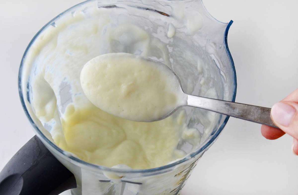 A person holding a spoon of creamy cauliflower soup over a blender containing more cauliflower soup.