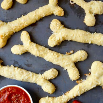 Several baked, cheesy breadstick bones with a small bowl of marinara sauce beside the breadsticks. An end of one of the breadsticks is dipped in marinara.