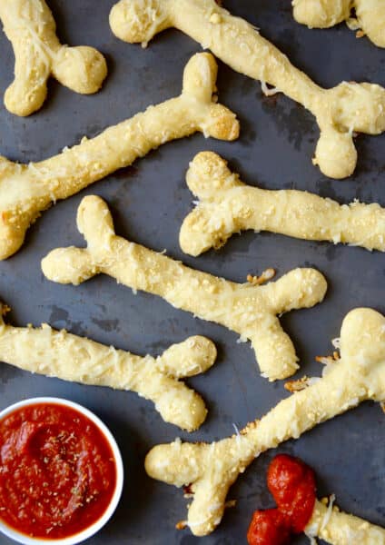 Several baked, cheesy breadstick bones with a small bowl of marinara sauce beside the breadsticks. An end of one of the breadsticks is dipped in marinara.