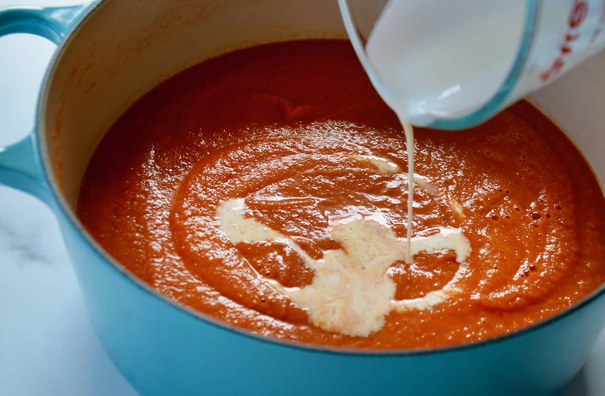 A glass measuring cup pours heavy cream into tomato soup in a light blue Dutch oven.