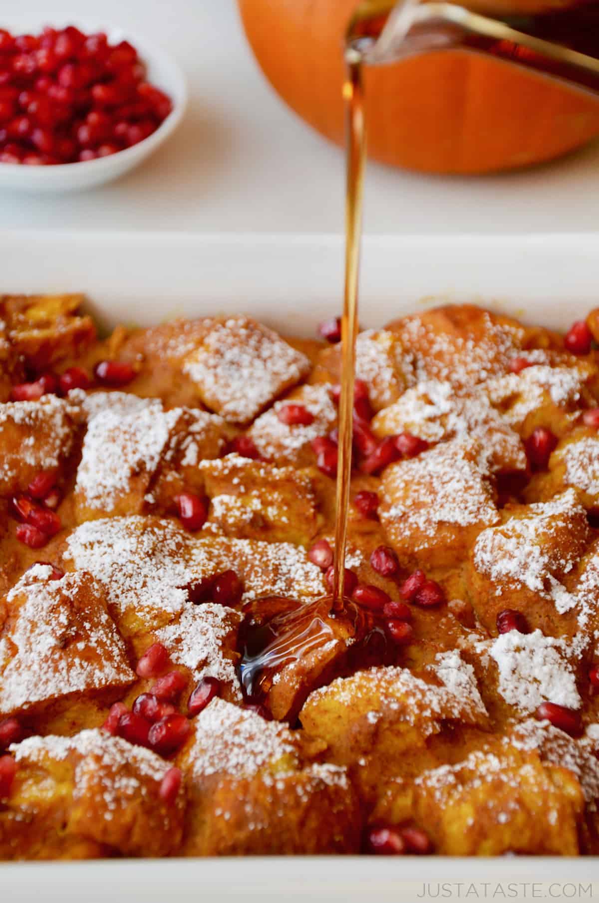 Maple syrup being poured onto a baking dish of pumpkin French toast casserole topped with pomegranate arils and powdered sugar.