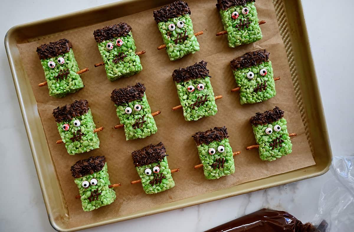 Three rows of Frankenstein Rice Krispie treats on a parchment-lined baking sheet. A piping bag filled with melted chocolate is beside the baking sheet.