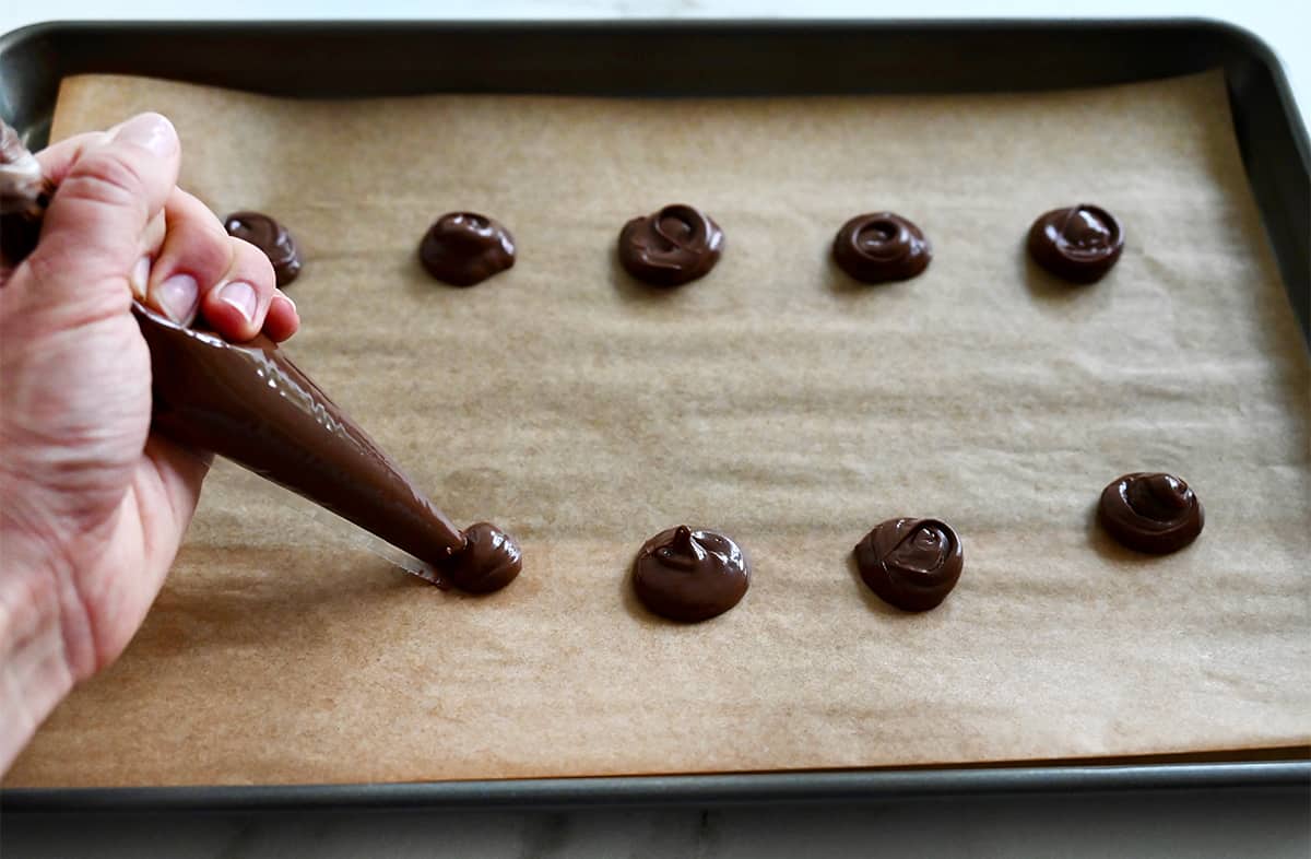 A hand with a piping bag makes small mounds of Nutella on a parchment paper-lined baking sheet.