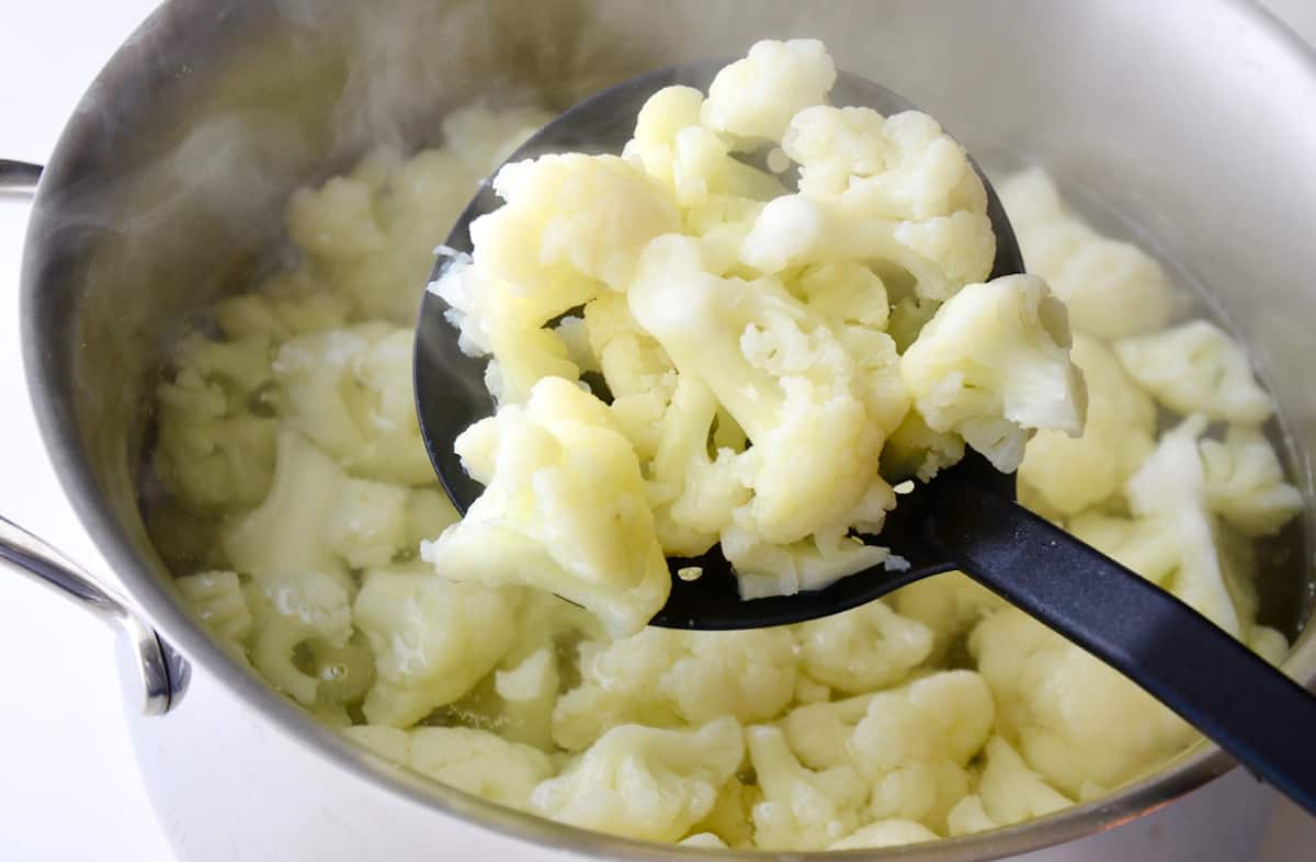 A slotted spoon holding cooked cauliflower florets over a large pot of boiling water containing more cooked cauliflower.