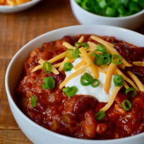 A bowl of turkey taco soup topped with sour cream, sliced scallions and grated cheese. Bowls of extra grated cheese and sliced scallions are beside it.