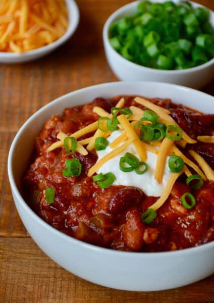 A bowl of turkey taco soup topped with sour cream, sliced scallions and grated cheese. Bowls of extra grated cheese and sliced scallions are beside it.