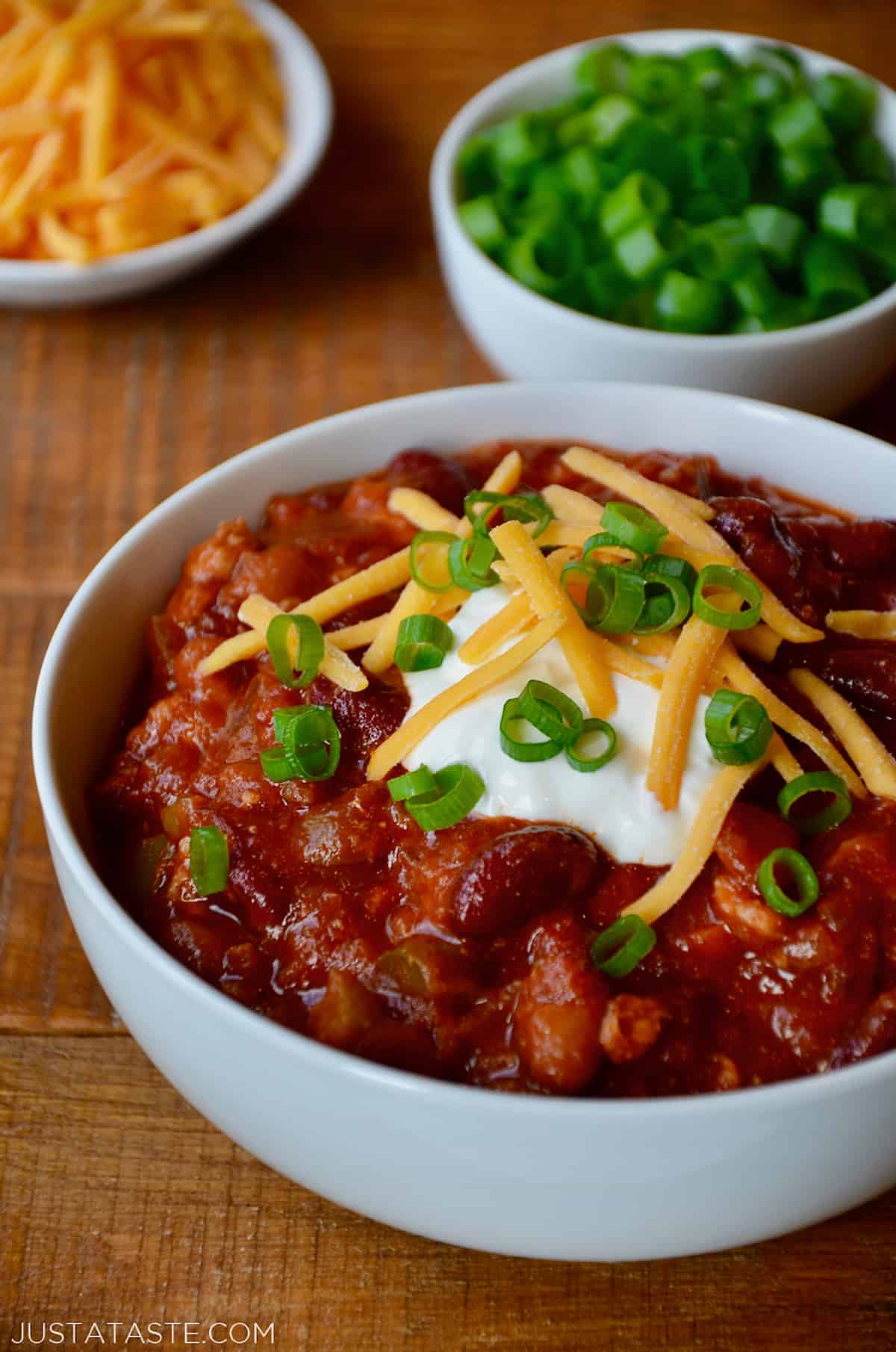 A bowl of turkey taco soup topped with sour cream, sliced scallions and grated cheese. Bowls of extra grated cheese and sliced scallions are beside it.