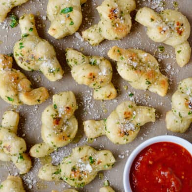 Garlic knots sprinkled with grated Parmesan cheese with a bowl of marinara sauce beside them.