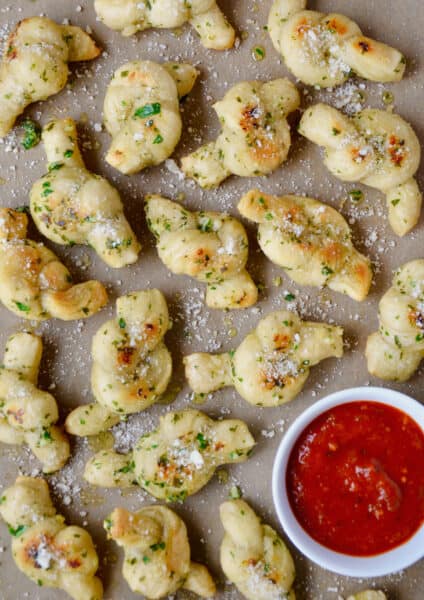 Garlic knots sprinkled with grated Parmesan cheese with a bowl of marinara sauce beside them.