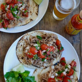 Two plates of carnitas tacos topped with cherry tomato pico de gallo and with cilantro and lime wedges to the side. A bottle of hot sauce and a beer glass sits beside the plates.