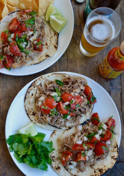 Two plates of carnitas tacos topped with cherry tomato pico de gallo and with cilantro and lime wedges to the side. A bottle of hot sauce and a beer glass sits beside the plates.