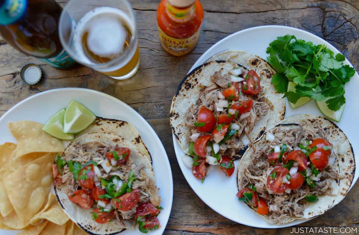 Two plates with Instant Pot Carnitas tacos topped with cherry tomato pico de gallo and with lime wedges, cilantro and tortilla chips on the plates next to them. A bottle of hot sauce and a glass with beer in it sits beside the plates.