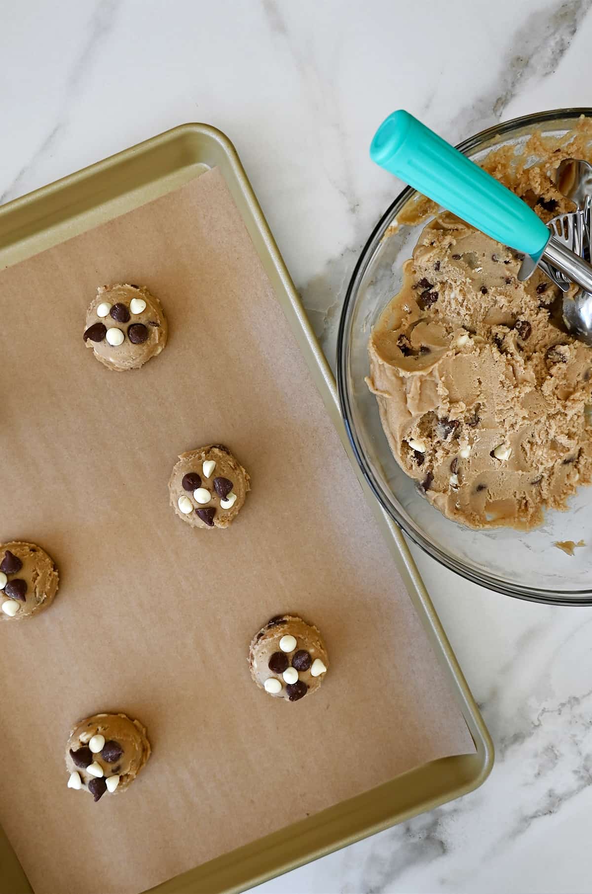 A bowl of cookie dough with an ice cream scoop next to a baking sheet with balls of cookie dough 