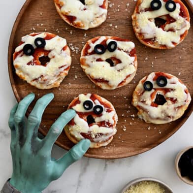 Six mummy mini pizza bagels on a wooden plate with small bowls of grated cheese and sliced olives beside the plate. A fake monster's hand is reaching over, pretending to grab a mini bagel.