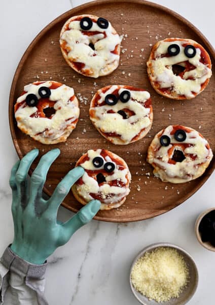Six mummy mini pizza bagels on a wooden plate with small bowls of grated cheese and sliced olives beside the plate. A fake monster's hand is reaching over, pretending to grab a mini bagel.