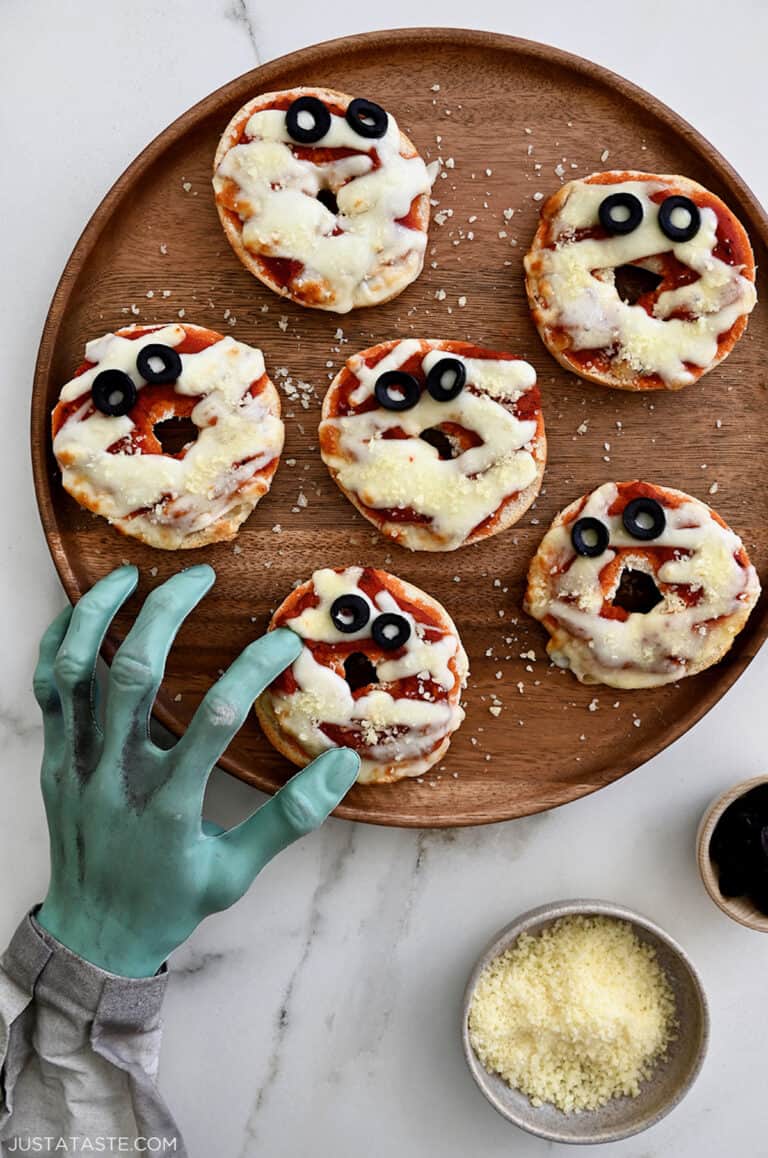 Six mummy mini pizza bagels on a wooden plate with small bowls of grated cheese and sliced olives beside the plate. A fake monster's hand is reaching over, pretending to grab a mini bagel.