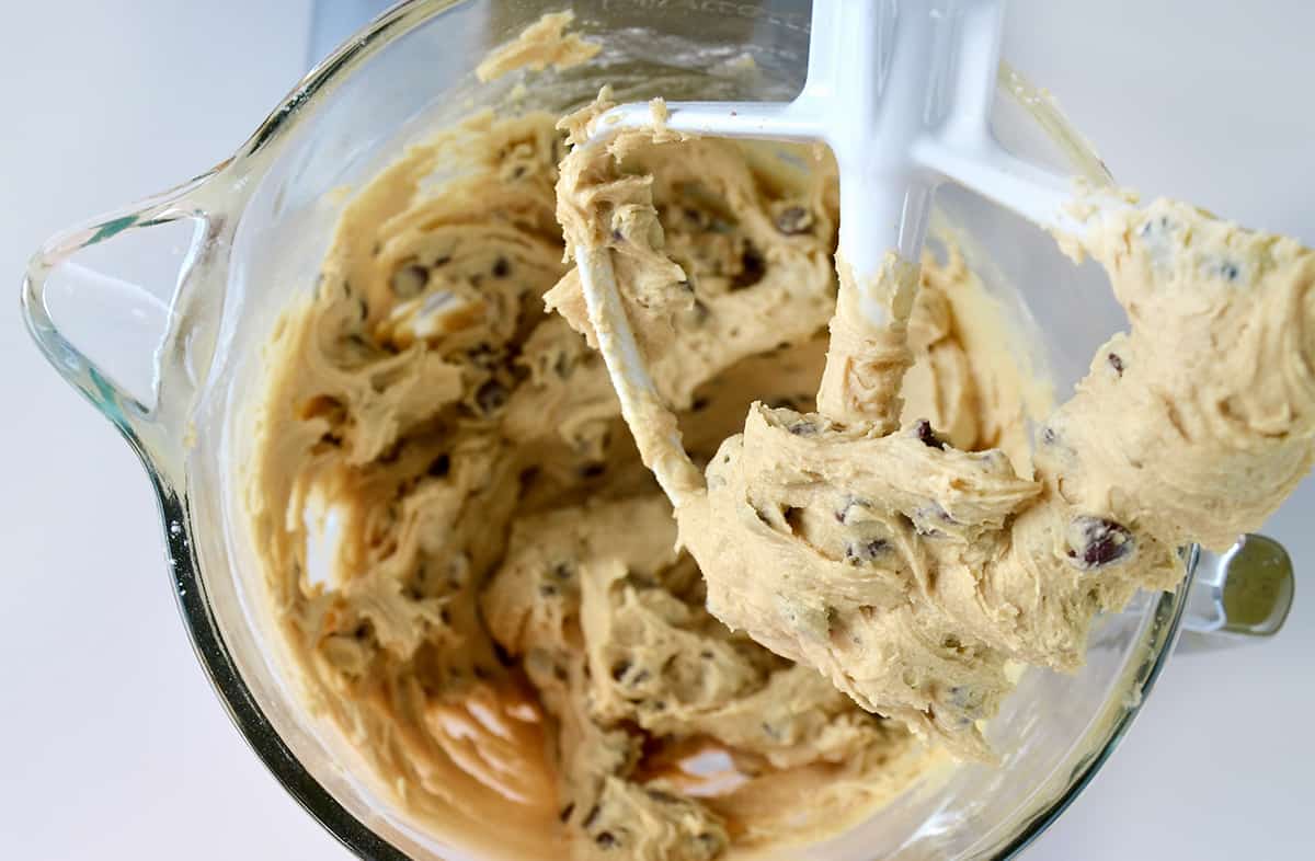 A paddle attachment of a stand mixer covered in chocolate chip cookie dough over a bowl containing more dough.