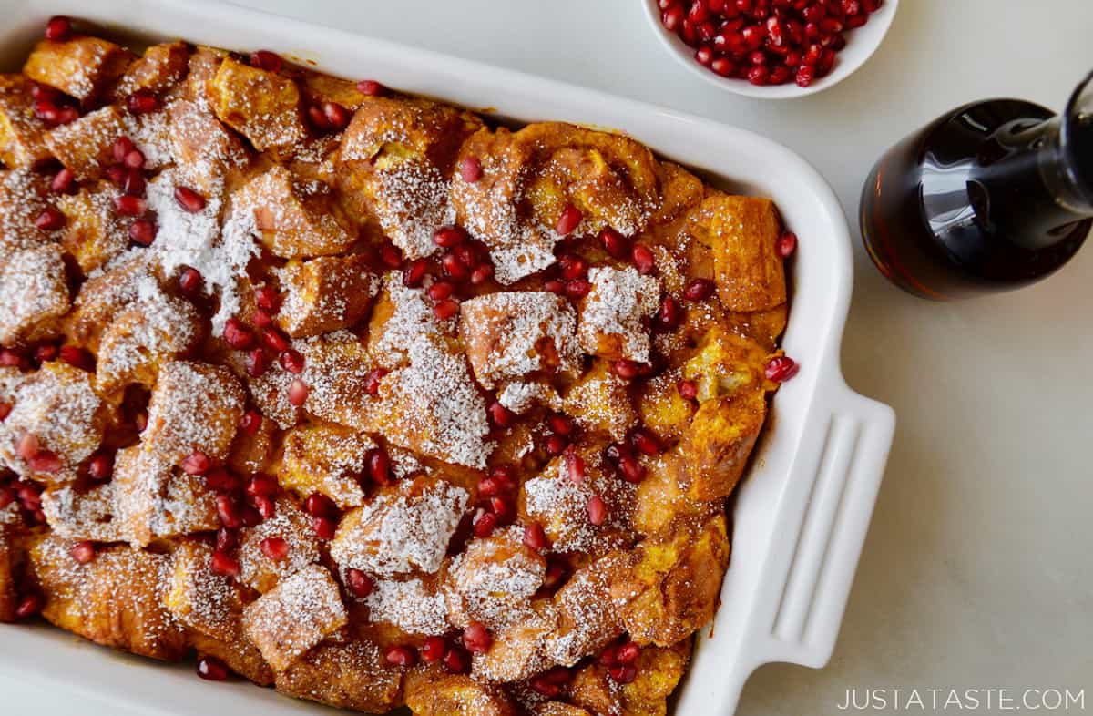 Baked pumpkin French toast in a baking dish that's topped with pomegranate arils and powdered sugar. Beside the dish is a carafe of maple syrup and a bowl of pomegranate seeds.