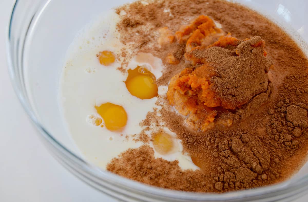 A glass mixing bowl with milk, eggs, pumpkin puree, sugar and pumpkin pie spice in it.
