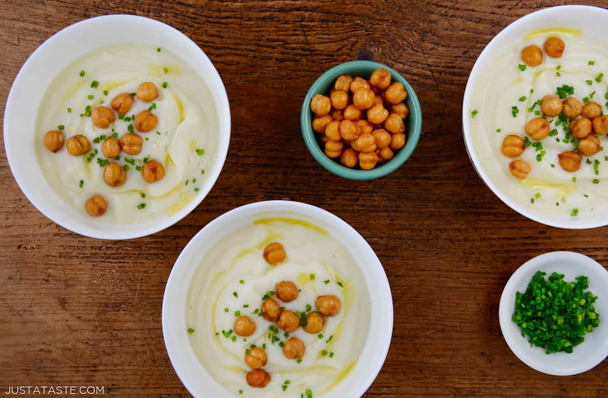 Three bowls of of creamy cauliflower soup topped with crispy chickpeas, minced chives and olive oil. Small bowls of extra chickpeas and minced chives are beside the bowls of soup.