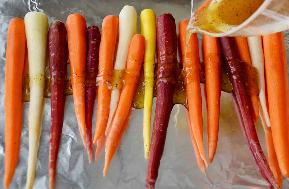 Peeled carrots on a foil-lined baking sheet. A mixture of honey, olive oil, salt and pepper is being poured over the carrots from a measuring cup.