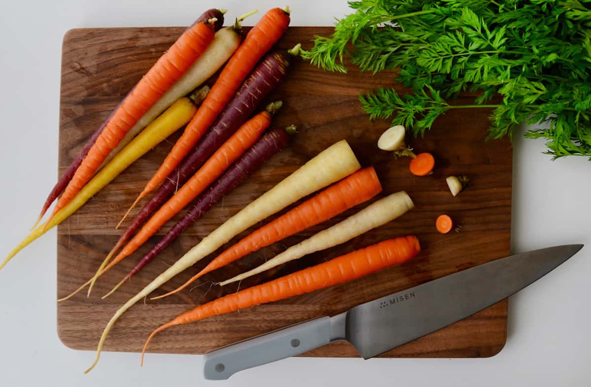 A group of rainbow carrots on a wooden cutting board with carrot tops and a chef's knife off to the side. Four off the carrots have some of their ends trimmed off.