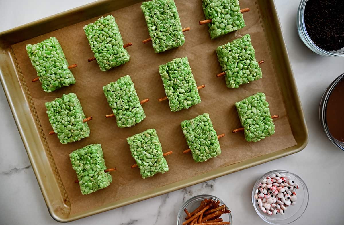 Green Rice Krispie treat rectangles with pretzel sticks stick out of their sides on a parchment-lined baking sheet. There are bowls of pretzel sticks, candy eyes, melted chocolate and candy sprinkles surrounding the baking sheet.