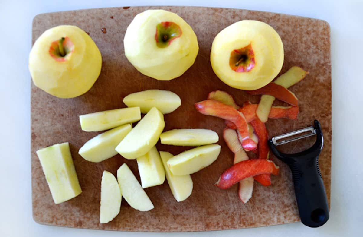 Three peeled whole apples and one peeled and sliced apple on a wooden cutting board. Apple peels and a vegetable peeler sit beside the apple slices.