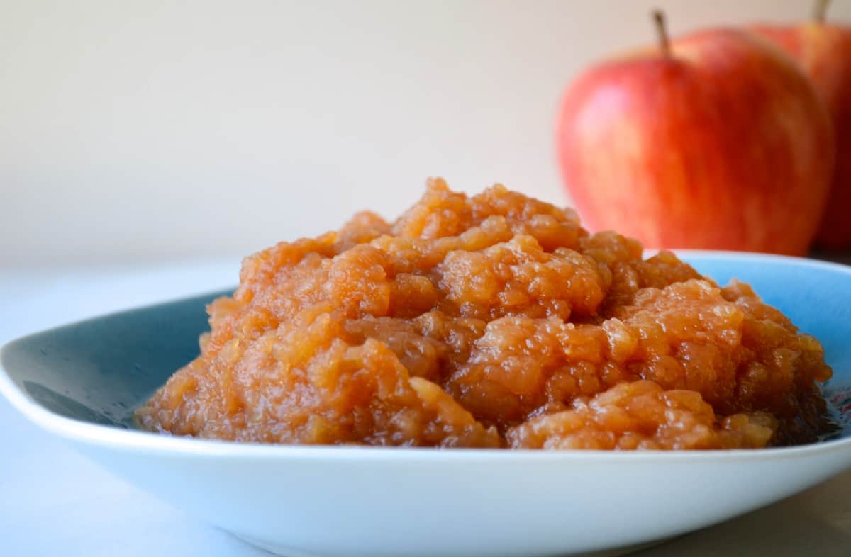 A blue bowl containing slow cooker applesauce. Two apples are stationed behind the bowl.