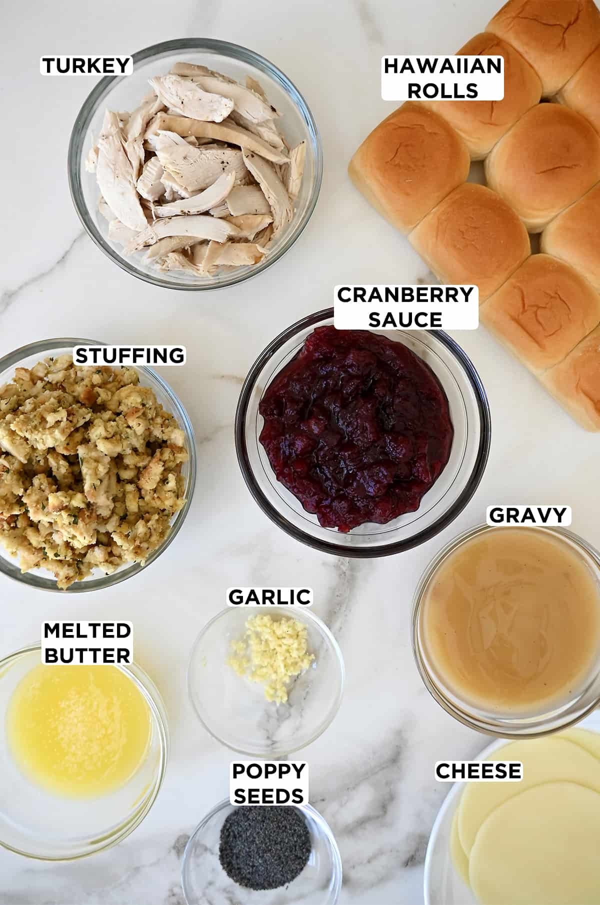 Hawaiian rolls next to various sizes of clear bowls containing shredded turkey, cranberry sauce, stuffing, gravy, garlic, melted butter, poppy seeds and slices of white cheese.