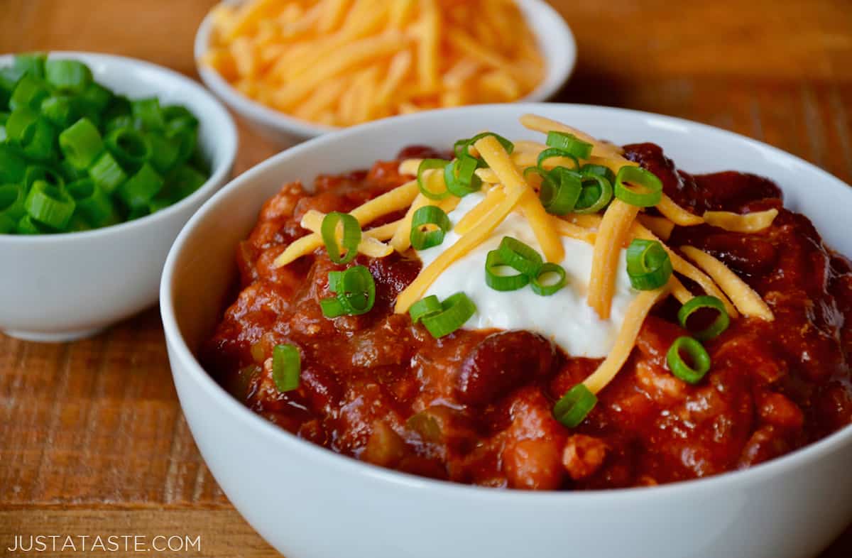A bowl of taco soup that's topped with sour cream, sliced scallions and grated cheese and has small bowls of extra grated cheese and sliced scallions beside it.