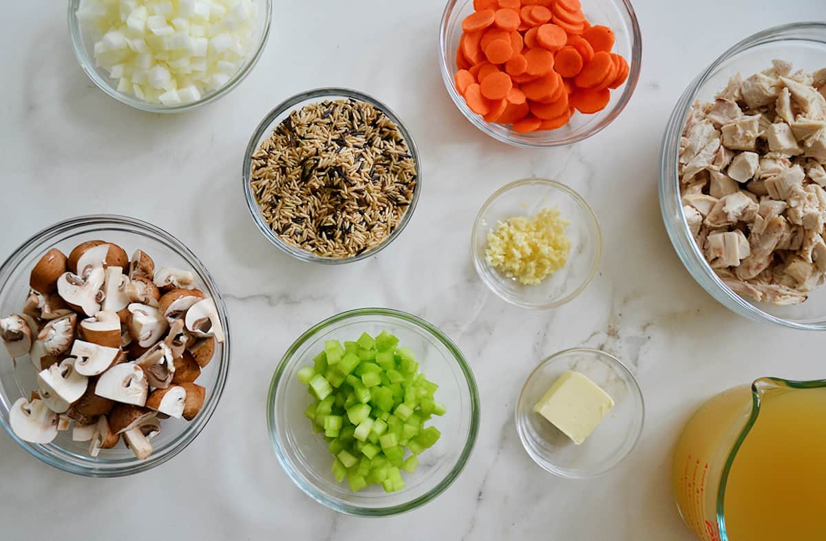 A glass measuring cup containing chicken broth along with several glass mixing bowls holding various ingredients (chopped onions, carrots and celery, minced garlic, butter, quartered mushrooms, wild rice and cooked chicken).