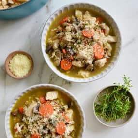 Two bowl of chicken wild rice soup topped with grated Parmesan cheese and thyme leaves. Surrounding the bowls are small bowls with cheese and thyme sprigs and a Dutch oven with the remainder of the soup in it.