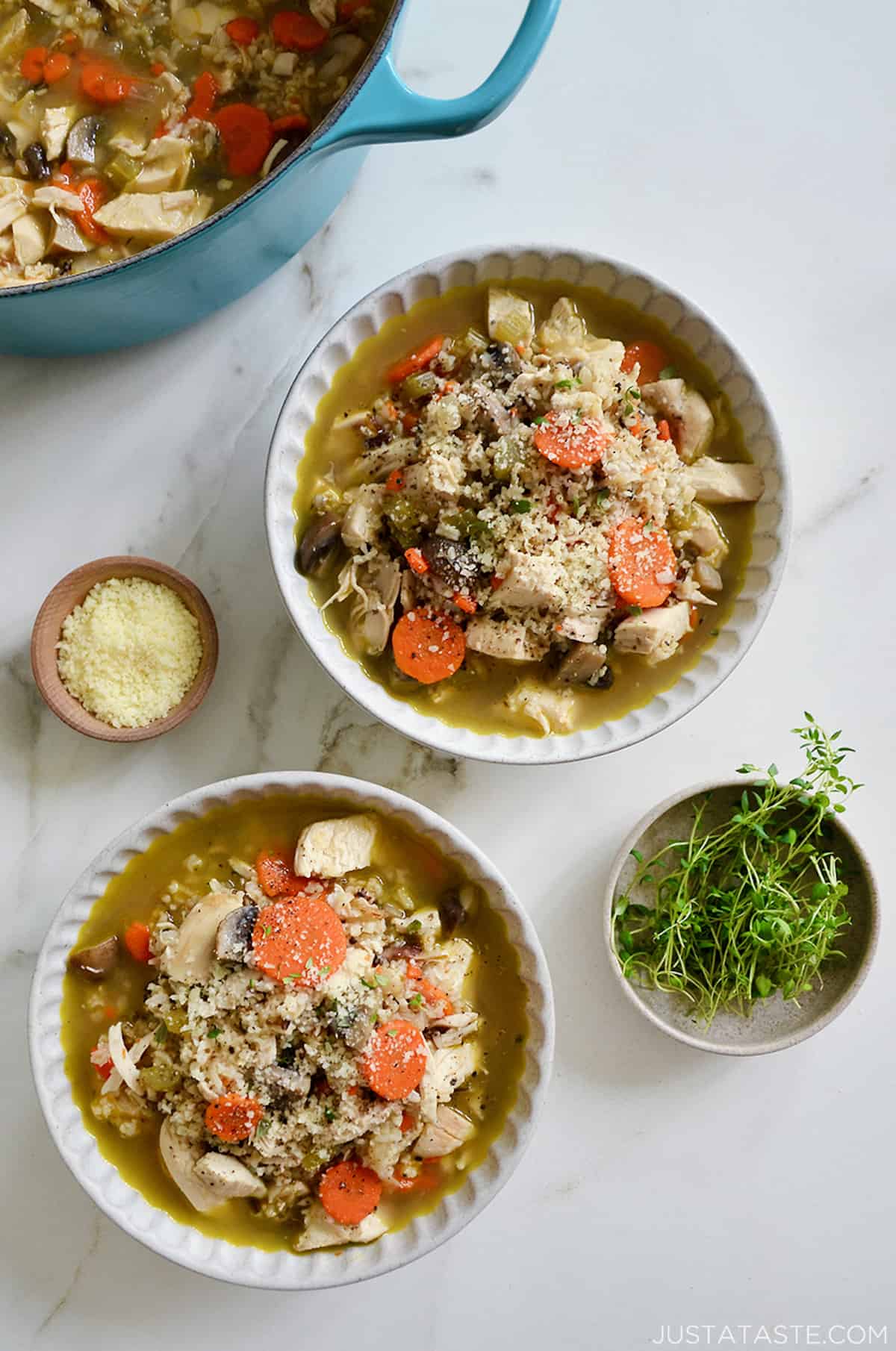 Two bowl of chicken wild rice soup topped with grated Parmesan cheese and thyme leaves. Surrounding the bowls are small bowls with cheese and thyme sprigs and a Dutch oven with the remainder of the soup in it.