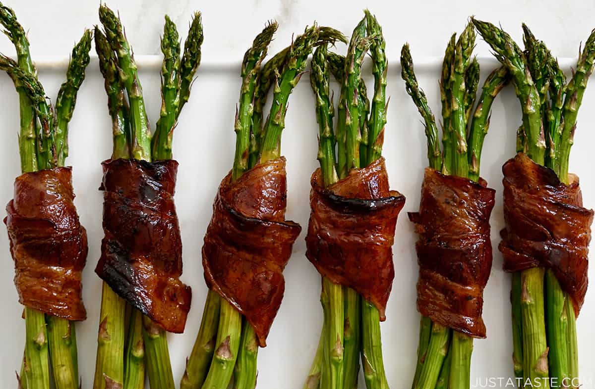 Six baked bacon-wrapped asparagus bundles on a white serving platter.